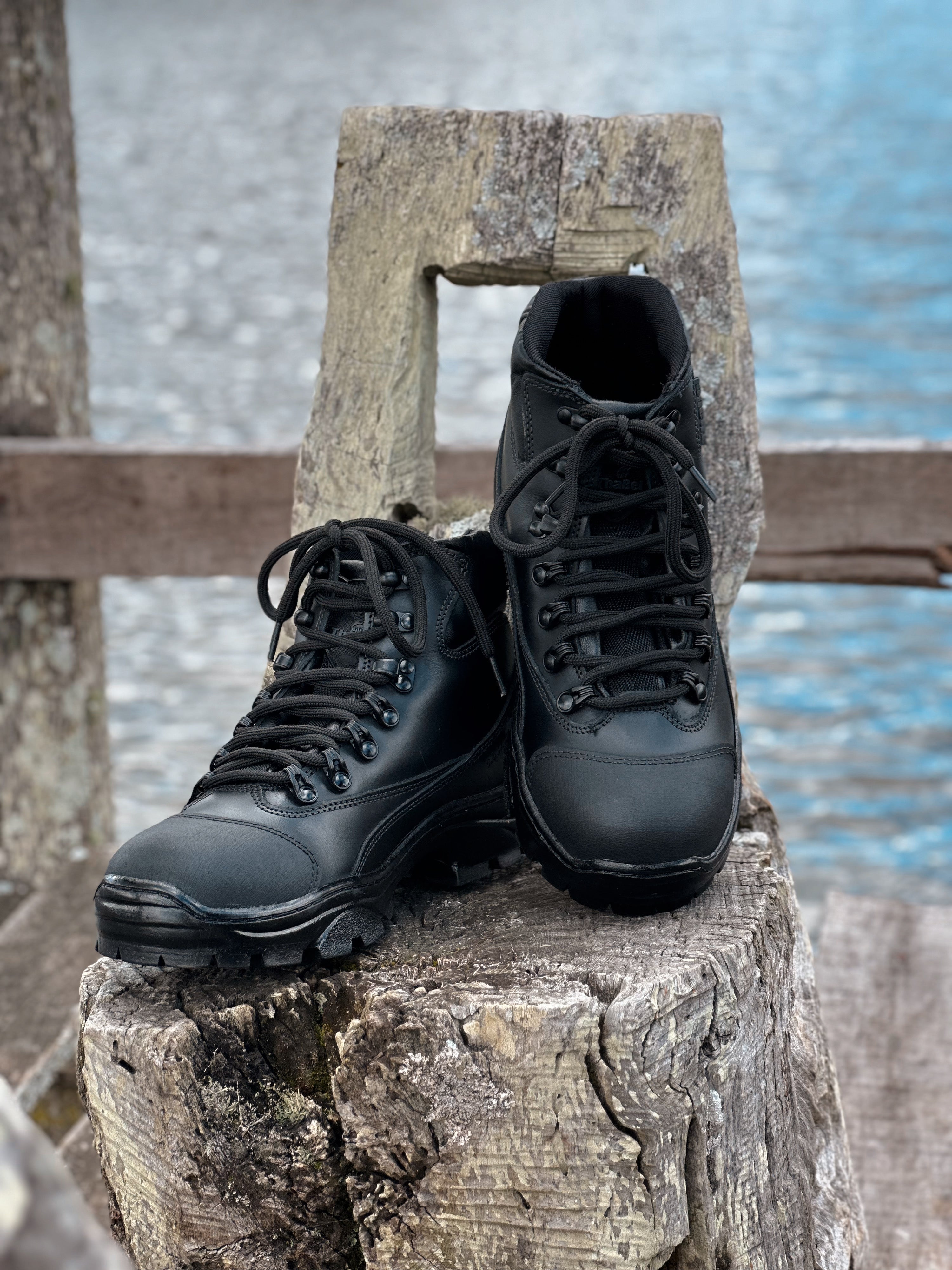 Bota cano médio em couro preta descansando sobre um tronco de madeira próximo à água.