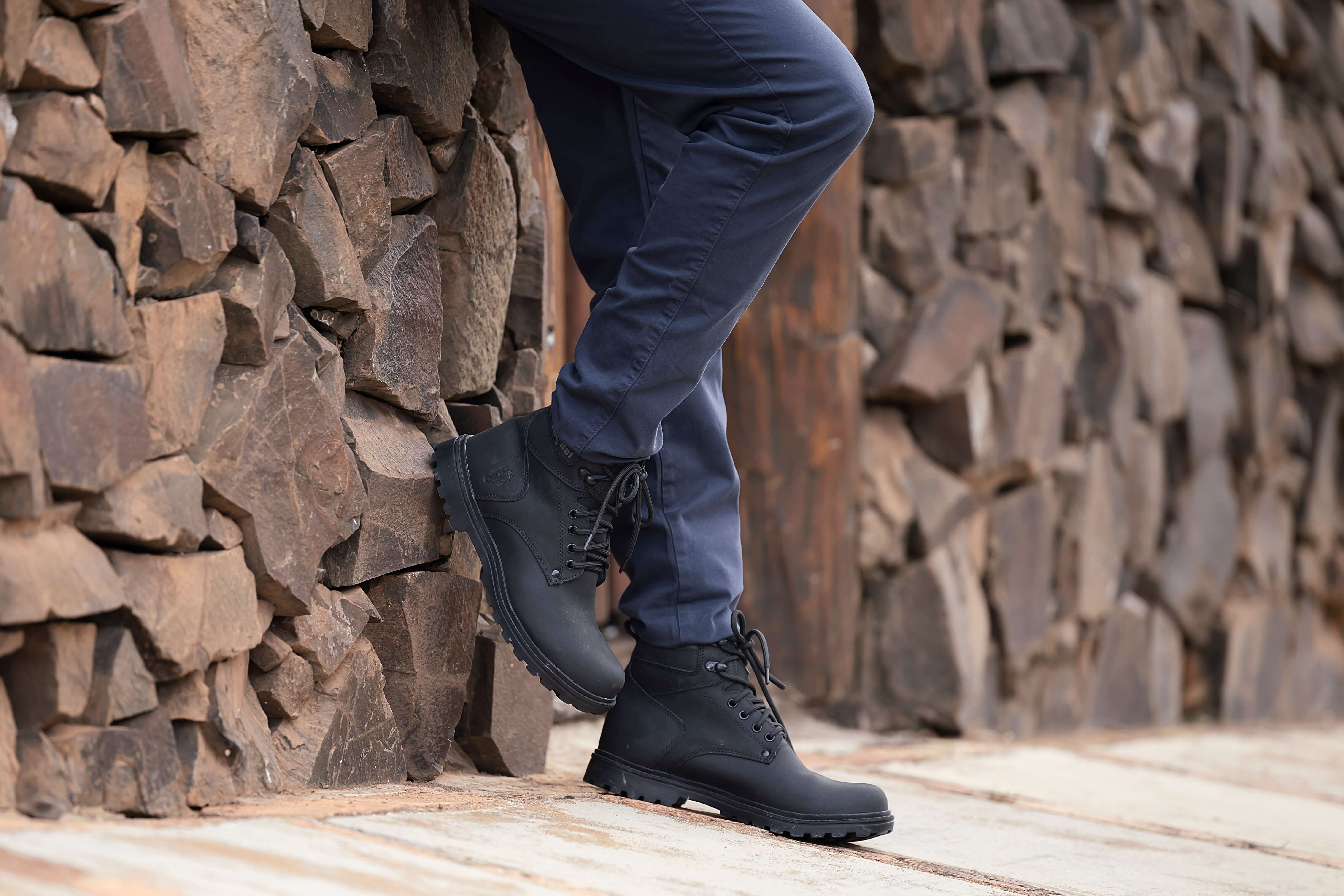 Pessoa usando calça azul e botinha masculina preta ao lado de uma parede de pedra. Botinha Masculina: Estilo, Tendências e Dicas de Uso.