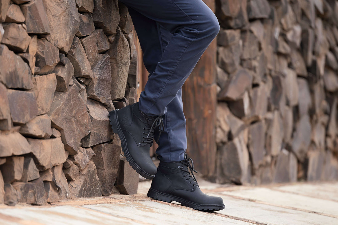 Pessoa usando calça azul e botinha masculina preta ao lado de uma parede de pedra. Botinha Masculina: Estilo, Tendências e Dicas de Uso.