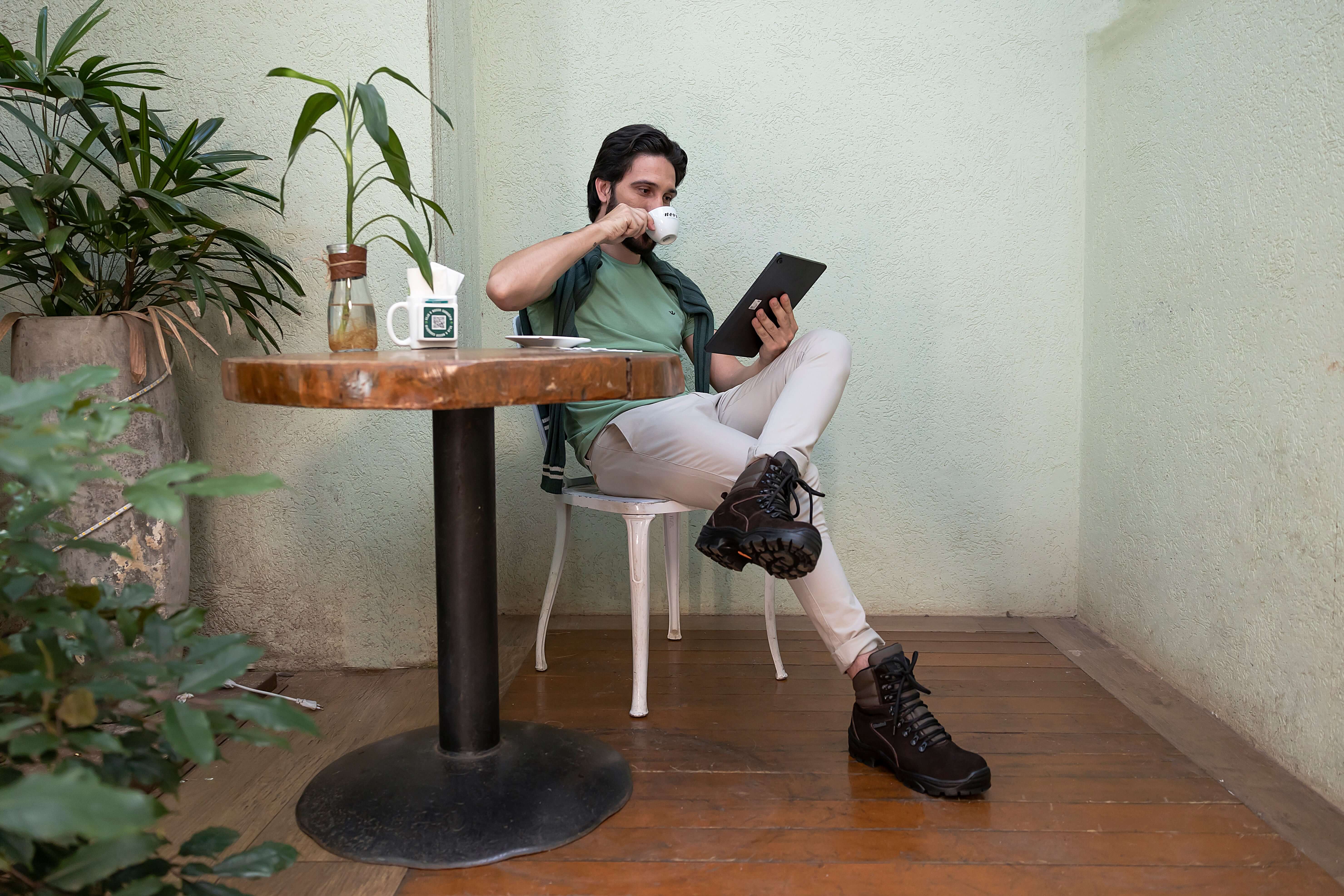 Homem sentado em um café usando Botas ThaBel enquanto lê, ilustrando "Descubra Por Que as Botas ThaBel São as Mais Indicadas".